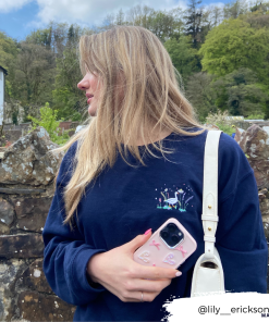 A person with long hair wearing a navy blue sweatshirt holds an iPhone15 encased in Coconut Lane's Pearly Hearts Phone Case - Pink and stands outdoors beside a stone wall. They have a white bag over their shoulder and are looking to the left, with trees and a building visible in the background. Instagram handle @lily__erickson is on the bottom right.