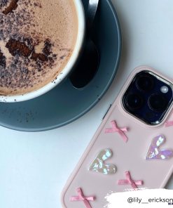 A cup of frothy hot chocolate in a white mug sits on a grey saucer with a spoon. Next to it is the Pearly Hearts Phone Case - Pink from Coconut Lane, decorated with pink bows and shiny heart stickers. The Instagram handle 