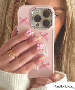 A person with long blonde hair partially covering their face holds a 'Pearly Hearts Phone Case - Pink' by Coconut Lane for an iPhone 15, decorated with pink bows and butterfly designs. Their manicured nails showcase pink bows, clear polish, and a butterfly design. They wear a cream-colored sweater. Text in the corner: "@naileditbeauty".