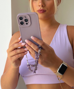 A person with blonde hair wearing a light purple sports bra from the NAKD spring collection takes a photo with a lavender-colored iPhone. They have light pink nails, and their phone is protected by the Coconut Lane NAKD Phone Case in Lilac, which features an amethyst beaded wrist strap for Full Camera Protection. They are also wearing a smartwatch on their left wrist.