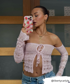 A woman takes a mirror selfie with a Wavy Phone Case - Berry Cute Strawberry from Coconut Lane. She wears a lacy, off-the-shoulder pink top with long sleeves tied at the front, paired with light blue jeans. Her hair is tied back, and she accessorizes with gold earrings and a necklace. The Instagram handle 