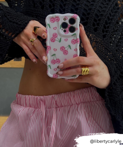 A person wearing a black crochet top and pink striped pants holds up a phone protected by the Wavy Phone Case - Disco Cherries from Coconut Lane. Their nails are adorned with nail art, and they are accessorized with several gold rings. The Instagram handle 