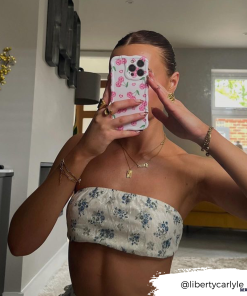 A person with slicked-back hair takes a mirror selfie, their face hidden by a phone adorned with Coconut Lane's Wavy Phone Case - Disco Cherries. They are dressed in a strapless, floral-patterned top. The room features modern decor and a large mirror. The Instagram handle @libertycarlyle_ is visible.