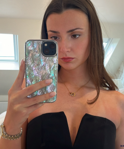 A young woman with long brown hair takes a mirror selfie inside a room with white walls and a window. She wears a strapless black top, a gold necklace with a crescent moon pendant, and a watch on her left wrist. Her phone case, the Real Natural Shell Phone Case in White by Coconut Lane, displays an iridescent, shell-like pattern.