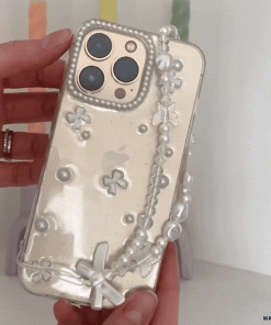 A hand is holding the Coconut Lane Mini White Bows Phone Case, adorned with a clear decorative design featuring pearl-like beads, small flower embellishments, and mini white bows. The background shows colorful, blurred objects.