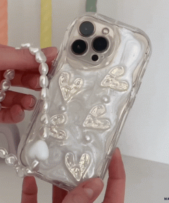 A person is holding the Pearly Hearts Phone Case - Clear by Coconut Lane, which features a pearlescent finish embellished with gold heart designs. This extra-protective iPhone 15 case also includes a detachable beaded strap resembling pearls. The background is a colorful blur of pastel hues.