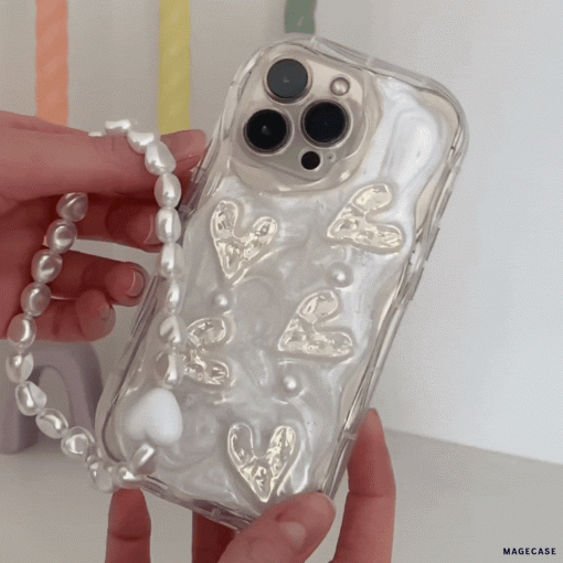 A person is holding the Pearly Hearts Phone Case - Clear by Coconut Lane, which features a pearlescent finish embellished with gold heart designs. This extra-protective iPhone 15 case also includes a detachable beaded strap resembling pearls. The background is a colorful blur of pastel hues.