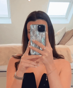 A woman with long dark hair is taking a mirror selfie using an iPhone protected by Coconut Lane’s Real Natural Shell Phone Case in White. She is dressed in a black top and minimal jewelry, including a bracelet and necklace. Behind her, you can see a neatly made bed and ceiling windows.