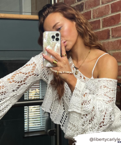 A woman, with her Instagram handle @libertycarlyle_, takes a mirror selfie with her phone encased in a Real Natural Shell Phone Case - White by Coconut Lane. She is wearing a white lacy top, as well as multiple rings and bracelets. Her long hair cascades over her shoulder while she gazes at the phone. The background features a brick wall and glass doors.