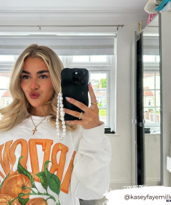 A woman with long blonde hair is taking a mirror selfie in a brightly lit room. She wears a white sweatshirt featuring a bold orange fruit design with the word 