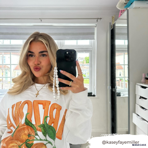 A woman with long blonde hair is taking a mirror selfie in a brightly lit room. She wears a white sweatshirt featuring a bold orange fruit design with the word "BRAVO" on it. Her phone, adorned with the Coconut Lane 3D Boujee Phone Case in an extra protective matte black finish, has a pearl chain attached. A dresser and shelves are visible in the background.