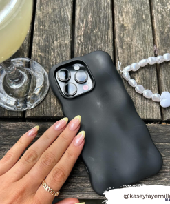A hand with manicured nails gently touches the Coconut Lane 3D Boujee Phone Case in matte black, displaying clearly visible camera lenses on a wooden surface. Beside it is a beaded bracelet with a white heart charm and a glass of light-colored beverage. Instagram handle @kaseyfayemiller.
