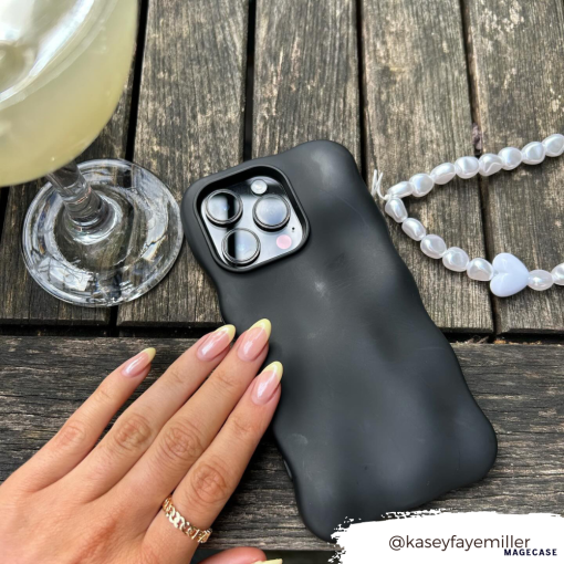 A hand with manicured nails gently touches the Coconut Lane 3D Boujee Phone Case in matte black, displaying clearly visible camera lenses on a wooden surface. Beside it is a beaded bracelet with a white heart charm and a glass of light-colored beverage. Instagram handle @kaseyfayemiller.