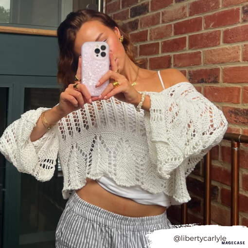 A person dressed in a loose white crochet sweater layered over a white top holds up an iPhone15 adorned with the Coconut Lane Moonstone Pink Phone Case, capturing a mirror selfie. They stand in front of a brick wall with part of a dark green door visible. The Instagram handle "@libertycarlyle_" is displayed at the bottom right.