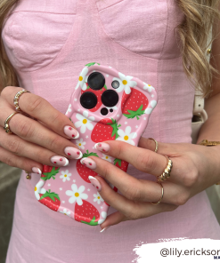 A person wearing a pink textured dress holds an extra protective Wavy Phone Case - Berry Cute Strawberry by Coconut Lane, adorned with strawberries and daisies. Their nails match the berry cute strawberry pattern. They are accessorized with multiple gold rings and a wristwatch. The text 