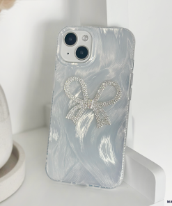 A close-up of a white iPhone with a dual-camera setup, encased in Coconut Lane's Diamante Bow Phone Case—a clear phone case adorned with a sparkly silver diamante bow design. The unique case not only adds flair but also provides extra protection. The phone is propped against a white surface, with a beige object partially visible in the background.