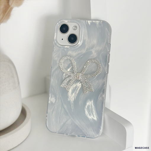 A close-up of a white iPhone with a dual-camera setup, encased in Coconut Lane's Diamante Bow Phone Case—a clear phone case adorned with a sparkly silver diamante bow design. The unique case not only adds flair but also provides extra protection. The phone is propped against a white surface, with a beige object partially visible in the background.