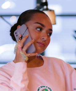 A person with a bun hairstyle is holding a phone in a NAKD Phone Case - Lilac by Coconut Lane to their ear, seemingly in conversation. They are wearing a light pink sweatshirt from the NAKD spring collection and standing indoors, with blurred background elements including lights and plants.