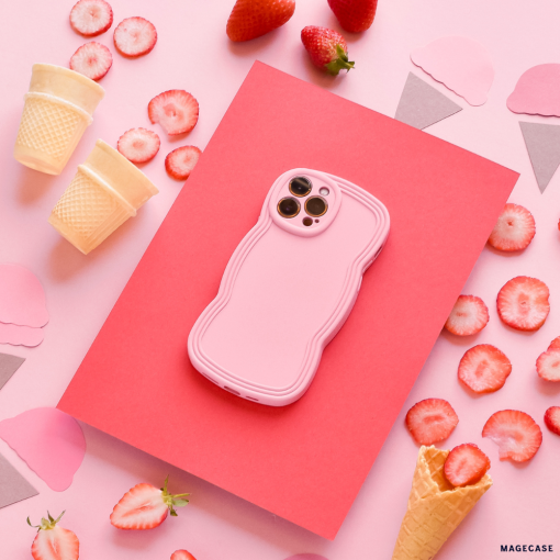 A pink smartphone encased in a Coconut Lane NAKD Curvy Phone Case - Strawberry, showcasing a textured, matte finish, is placed on a red sheet against a pink background. Surrounding it are sliced strawberries, whole strawberries, ice cream cones, and cutouts of ice cream shapes, creating a colorful dessert-themed arrangement.