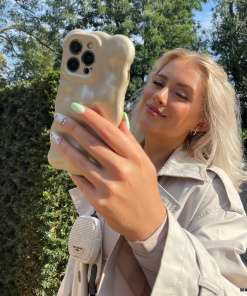 A woman with long blonde hair and mint green nails takes a selfie outdoors. She is wearing a beige trench coat and smiling softly. Her iPhone 15 is adorned with Coconut Lane’s Gold 3D Pearl Phone Case, shaped like a bear, and a small white device is attached to her bag strap. Trees and a hedge are in the background.