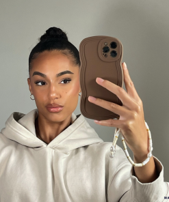 A woman with her hair in a high bun takes a selfie in front of a mirror. She is wearing a light beige hoodie and holding a brown phone with the lightweight, highly protective NAKD Curvy Phone Case in 