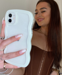 A young woman with long brown hair smiles while taking a selfie. She wears a brown, strapless top and has multiple rings on her fingers. Her iPhone case by Coconut Lane, the Curvy Phone Case in Cream, notably a highly protective design with sleek curves, stands out against the plain background.
