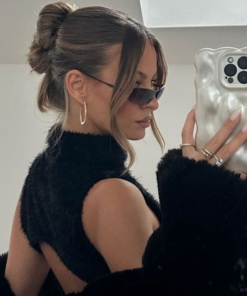 A woman with her hair in an elegant bun wears sunglasses, hoop earrings, and a black sleeveless top. She is taking a mirror selfie with her iPhone encased in the Coconut Lane 3D Pearl Phone Case - White, showing off her long nails and rings on her fingers. The background is an indoor setting.