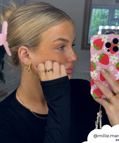 A person with blonde hair held back by a pink hair clip is taking a selfie in a mirror. They are wearing gold rings and earrings, a black top, and holding an extra protective phone encased in the Wavy Phone Case - Berry Cute Strawberry from Coconut Lane. The Instagram handle 