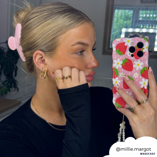 A person with blonde hair held back by a pink hair clip is taking a selfie in a mirror. They are wearing gold rings and earrings, a black top, and holding an extra protective phone encased in the Wavy Phone Case - Berry Cute Strawberry from Coconut Lane. The Instagram handle "@millie.margot" is shown in the image.