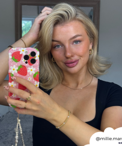 A woman with shoulder-length blonde hair poses for a mirror selfie. She holds her Coconut Lane Wavy Phone Case - Berry Cute Strawberry. She wears gold jewelry and a black top. The Instagram handle 