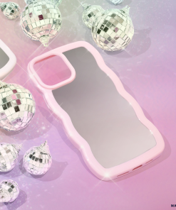 A pink, wavy-edge phone case lies on a pastel, shimmering background. Surrounding the Curvy Mirror Phone Case - Pink by Coconut Lane are small, reflective disco ball ornaments, adding a festive and sparkly touch to the scene. The background blends hues of purple and pink, accentuating the glittery ambiance.