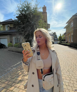 A woman with long blonde hair stands on a cobblestone driveway between two large houses under a bright, sunny sky. She is wearing a light-colored trench coat, a crop top, and ripped jeans. In one hand, she holds her iPhone15 adorned with Coconut Lane's 3D Pearl Phone Case - Gold and carries two small crossbody bags.