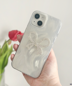 A hand holding a smartphone featuring the Diamante Bow Phone Case by Coconut Lane, an extra protective, pearlized white case adorned with a sparkly diamante bow design. In the blurred background, there's a red tulip in a clear vase.