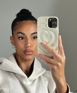 A person with neatly styled hair in a bun takes a selfie using a smartphone adorned with Coconut Lane's Melting Heart Phone Case in Coconut Cream. They are wearing a light-colored hoodie and gold hoop earrings, maintaining a neutral facial expression against a plain background.