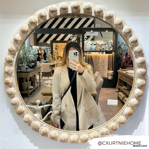 A person wearing a beige blazer and black top takes a mirror selfie in a home decor store. Various items like furniture and decorative pieces are displayed in the background. The mirror has a round, ornate frame with a beaded design. They're holding the Curvy Phone Case - Cream by Coconut Lane, which is highly protective for their iPhone 15. Text in the corner reads "@CXURTNIEHOME.
