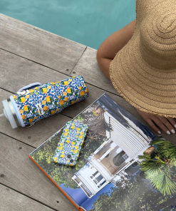 A person wearing a straw hat is lying beside a pool with an Amalfi Lemon-patterned tumbler, a Coconut Lane Wavy Phone Case - Amalfi Lemon, and a book featuring photographs of a house. The person has manicured white nails and is holding the book open to a page displaying an image of a white house.