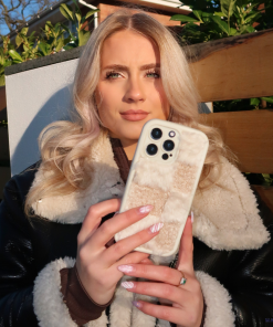 A young woman stands outdoors holding up a Coconut Lane Cosy Teddy Phone Case in Vanilla Biscuit. She has long, wavy blonde hair and is wearing a black jacket with a fluffy, light-colored inner lining. The background shows greenery and a brown wooden fence.