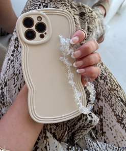A person holds a beige smartphone with the NAKD Curvy Phone Case - Vanilla by Coconut Lane, featuring a unique wavy-edged design and paired with a clear beaded strap. The person's nails are manicured in light pink, and they are wearing a snake-print patterned outfit. A gold bracelet adorns their wrist, perfectly complementing the highly protective yet lightweight phone case from Coconut Lane.