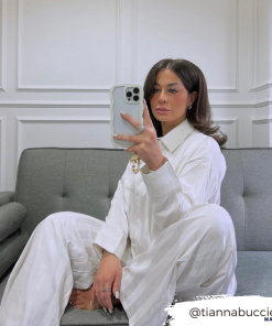 A woman with long dark hair is sitting on a gray couch taking a mirror selfie with her phone. She is wearing a white striped pajama set, and her Curvy Phone Case - Cream from Coconut Lane shines in the shot. The background features a white paneled wall. The Instagram handle 