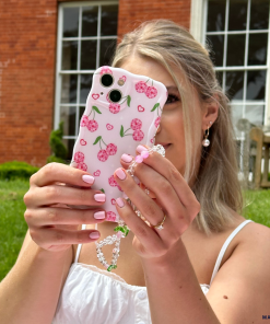 A person with long, light-colored hair is holding a smartphone encased in Coconut Lane's Wavy Phone Case - Disco Cherries, complete with an attached beaded bracelet. She is taking a photo outdoors in front of a building with large windows and a grassy lawn, showcasing her pearl earring and manicured nails.