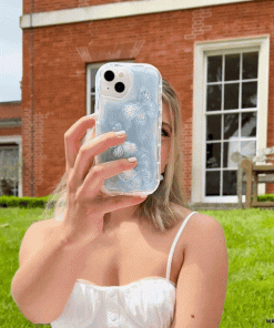 A woman in a white sleeveless dress takes a selfie with a phone encased in the Coconut Lane Shimmer Blue Butterflies Phone Case. She is outdoors in front of a brick building with large windows and green grass.