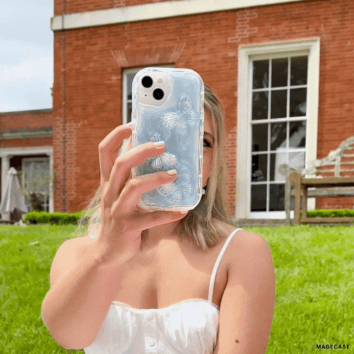 A woman in a white sleeveless dress takes a selfie with a phone encased in the Coconut Lane Shimmer Blue Butterflies Phone Case. She is outdoors in front of a brick building with large windows and green grass.