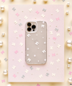 A smartphone with a Coconut Lane Mini White Bows Phone Case featuring a transparent floral and pearl design is placed on pink paper, adorned with small shiny decorations. The backdrop showcases a pastel color scheme with scattered pearls, pink stars, and glittery silver leaf shapes.