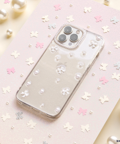 A smartphone adorned with the Mini White Bows Phone Case from Coconut Lane rests on a glittery pastel background, enhanced by scattered pink and silver star-shaped confetti and pearls. The phone’s triple camera setup is elegantly visible, contributing to a whimsical and sophisticated aesthetic.