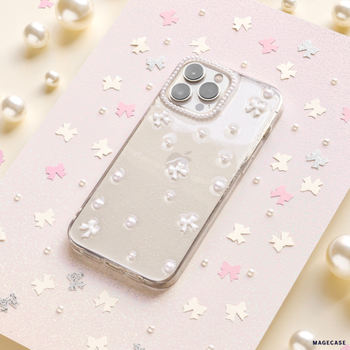 A smartphone adorned with the Mini White Bows Phone Case from Coconut Lane rests on a glittery pastel background, enhanced by scattered pink and silver star-shaped confetti and pearls. The phone’s triple camera setup is elegantly visible, contributing to a whimsical and sophisticated aesthetic.