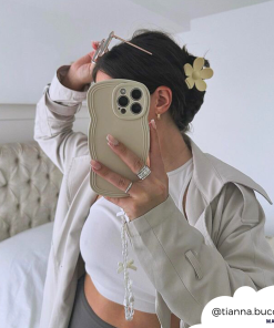 Person takes a mirror selfie with an iPhone in a highly protective, beige wavy case by Coconut Lane's NAKD Curvy Phone Case in Vanilla. They're wearing a cream jacket and white crop top, their hair is up with a flower clip. A white bed and minimal decor are in the background. Instagram handle 