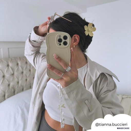 Person takes a mirror selfie with an iPhone in a highly protective, beige wavy case by Coconut Lane's NAKD Curvy Phone Case in Vanilla. They're wearing a cream jacket and white crop top, their hair is up with a flower clip. A white bed and minimal decor are in the background. Instagram handle "@tianna.buccieri" is displayed.