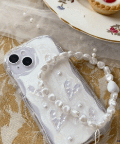 A white iPhone with a Coconut Lane Pearly Hearts Phone Case - Clear, showcasing pearl-like accents and transparent heart shapes, rests on an ornate gold-patterned tablecloth. A white bracelet with pearl-like beads lies atop the phone. In the background, there is a floral-patterned dessert plate filled with pastries.