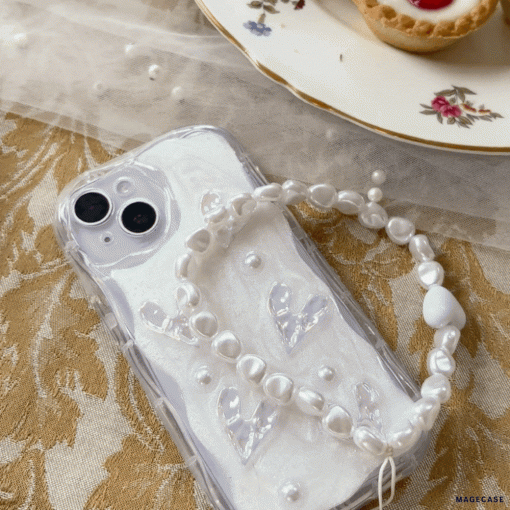 A white iPhone with a Coconut Lane Pearly Hearts Phone Case - Clear, showcasing pearl-like accents and transparent heart shapes, rests on an ornate gold-patterned tablecloth. A white bracelet with pearl-like beads lies atop the phone. In the background, there is a floral-patterned dessert plate filled with pastries.