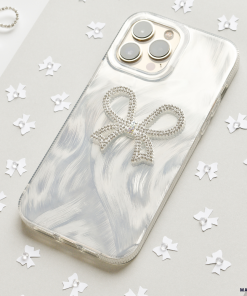 A smartphone encased in the Coconut Lane Diamante Bow Phone Case, which boasts a sparkling bow decoration, rests on a light gray surface. This distinctive phone case infuses an element of elegance, while the background is sprinkled with scattered white, star-like confetti. The phone's camera lenses are clearly visible, prominently situated at the top left corner.